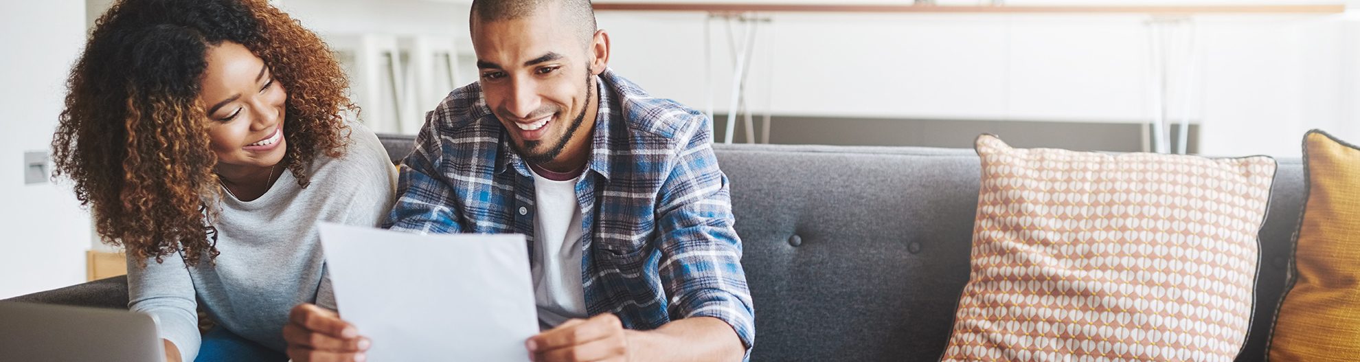 Image of young couple sitting on the couch reading the paper and sitting in front of their laptop